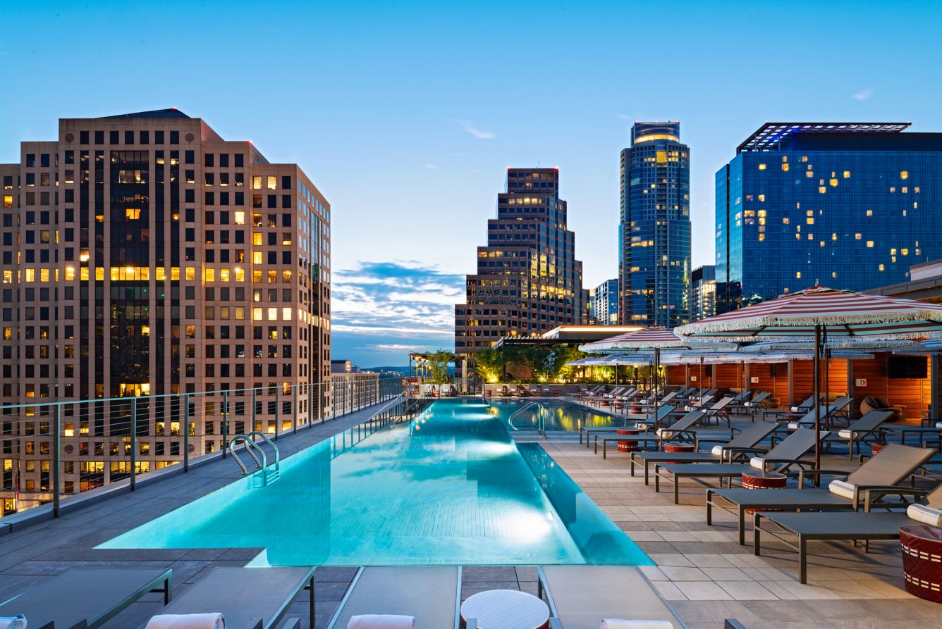 Rooftop pool at dusk, with city skyscrapers glowing in the background and poolside lounge chairs under striped umbrellas creating a luxurious urban retreat.